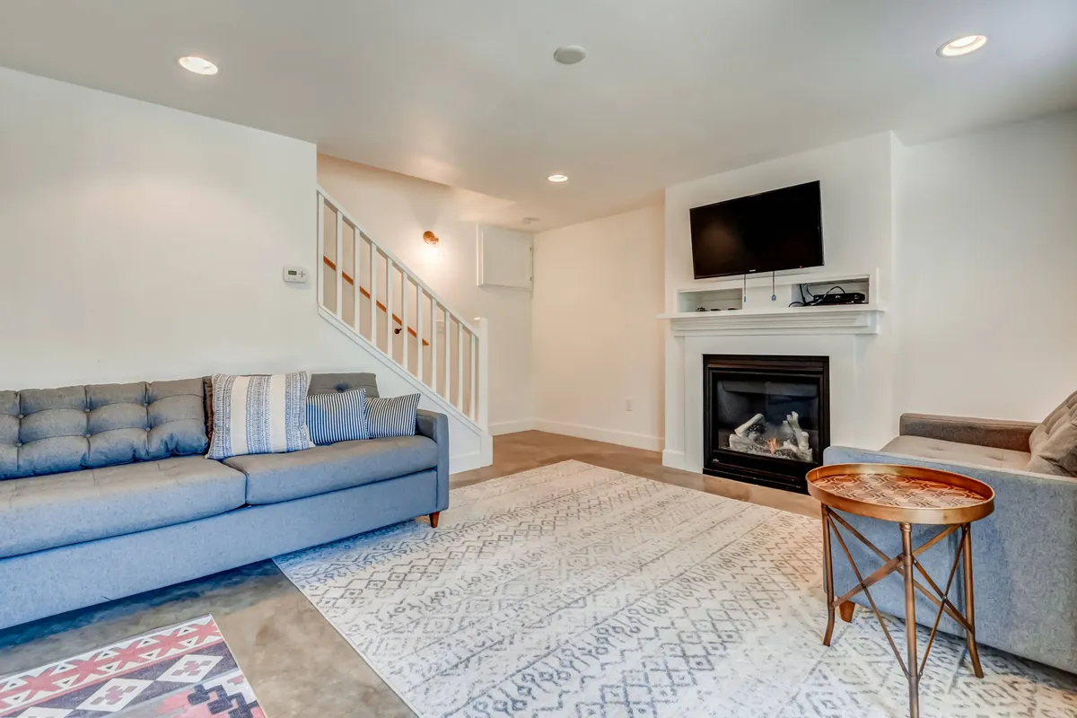 A cozy living room featuring a comfy sofa, modern fireplace, large television, textured rug, and an inviting staircase. The design blends warmth with style, making it a natural gathering spot. The room is bathed in natural light, highlighting both the bohemian decor and magazine-worthy finishes.