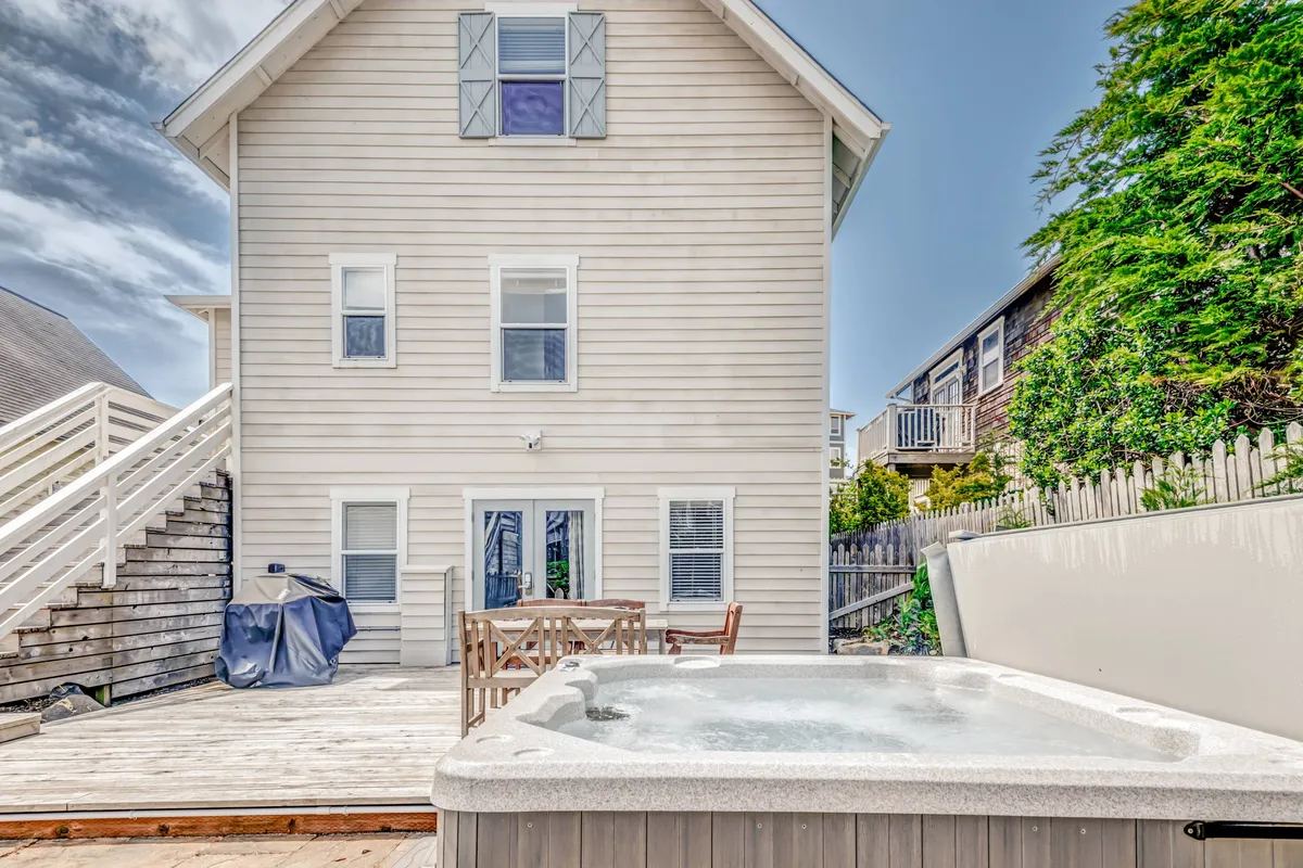 This image features a spacious backyard deck with a bubbling hot tub, comfortable outdoor seating, and stairs leading to an upper level. The area is designed for both relaxation and socializing, with views of the greenery beyond the deck.