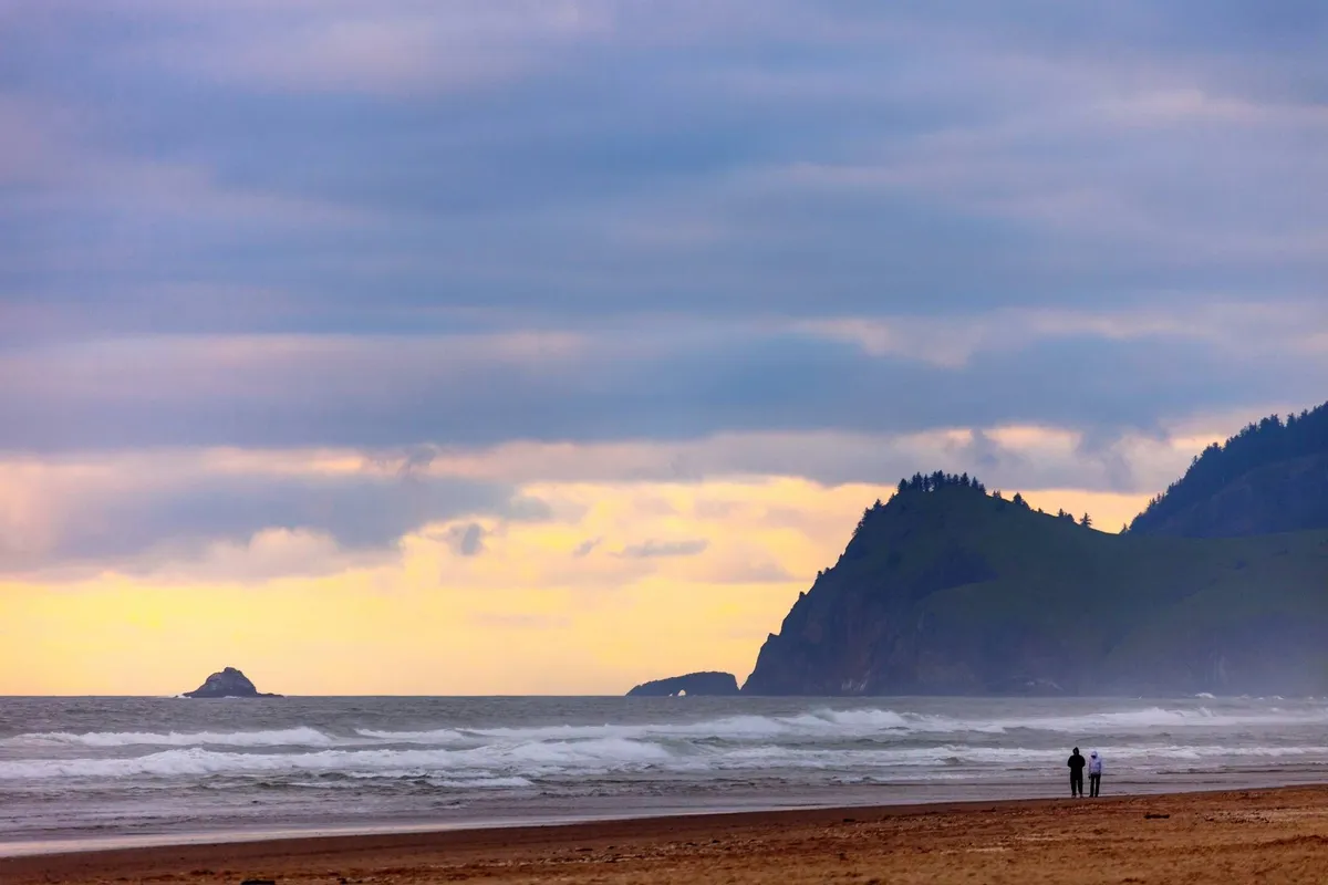 This image captures a wide view of Lincoln City Beach, with waves rolling onto sandy shores, distant cliffs framing the coastline, and a couple taking a stroll along the water's edge. The sun is low, casting a golden glow that highlights the natural beauty of the area.
