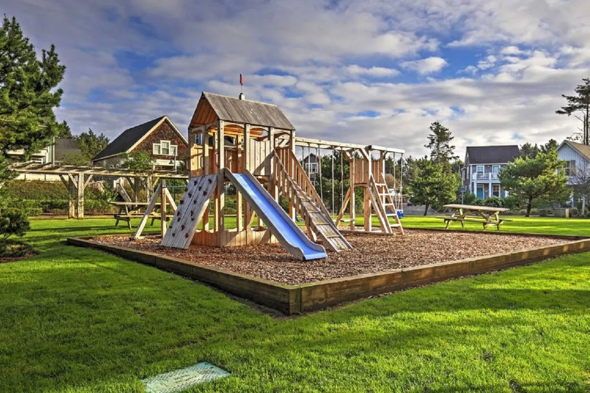 A vibrant community playground surrounded by green lawns, with swings, slides, and picnic tables. Children are playing and families are gathered, suggesting a joyful, family-friendly atmosphere.