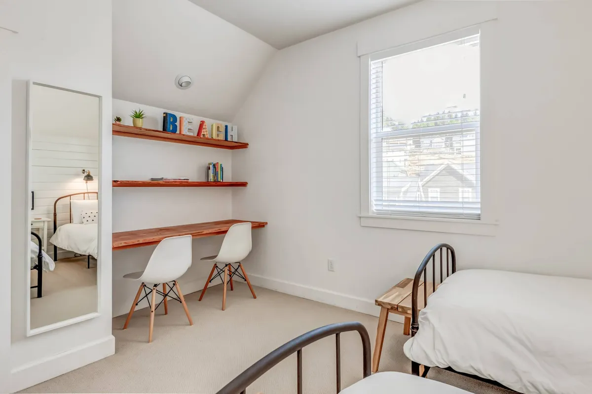 A calm, inviting bedroom with a comfortable bed, modern desk and chairs, shelves displaying books and thoughtful décor, and a large mirror. The overall feel is light and tranquil, with ample space for both rest and focus.