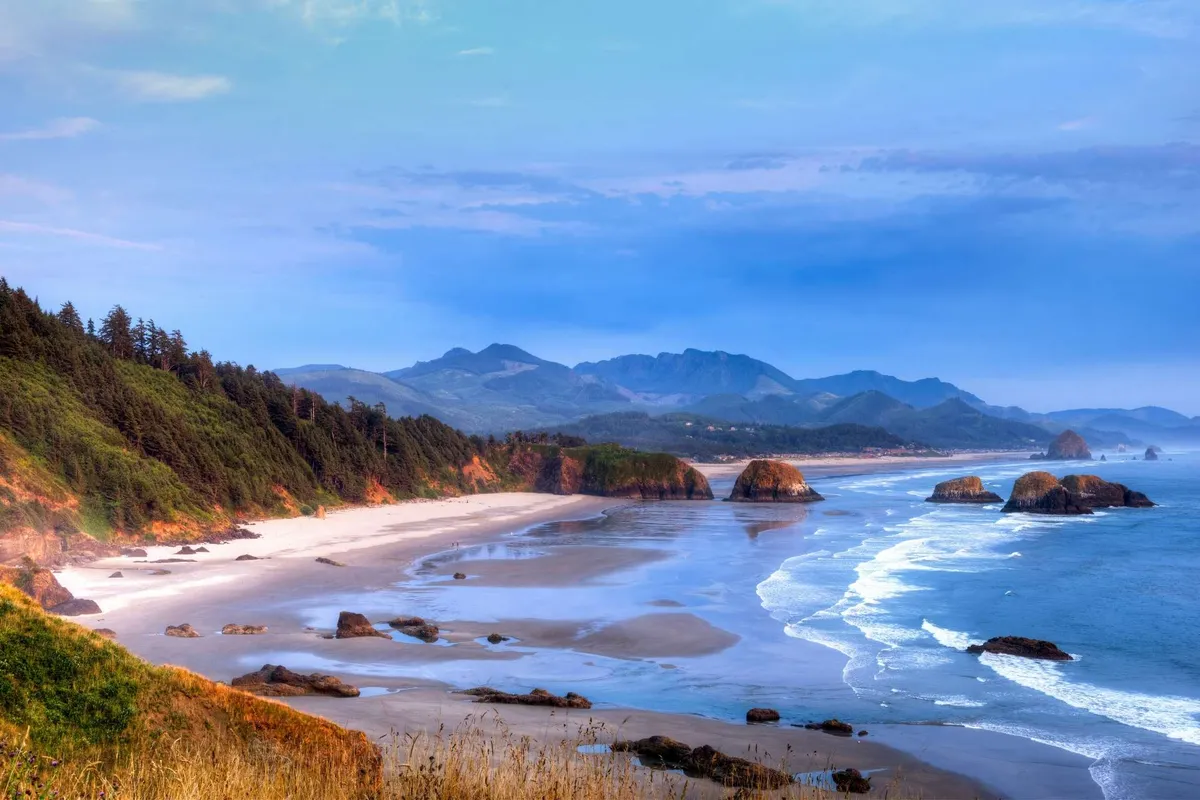 A panoramic view of the coastline featuring distant mountains, a rocky shoreline, sandy beach, and gentle ocean waves. The landscape is tranquil and inviting, ideal for exploring, beachcombing, or relaxing while taking in the stunning scenery.