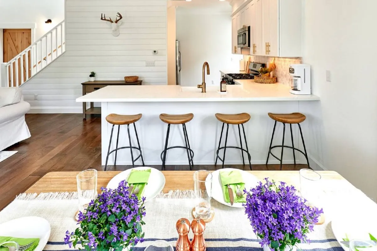 A focused interior shot showcasing the kitchen island with stools, a dining table adorned with fresh flowers, and plenty of light filtering through the windows. The layout emphasizes the accessible, central nature of the kitchen and its role as a hub for cooking, dining, and conversation.