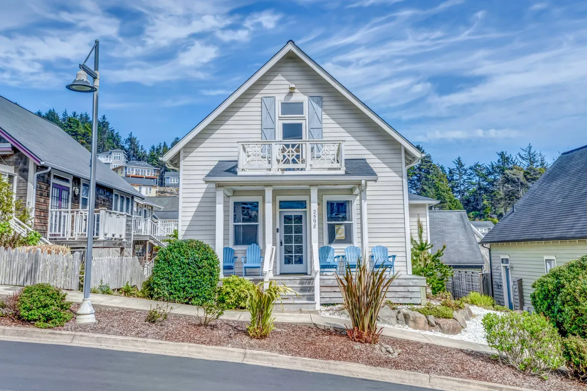 A welcoming front porch with inviting blue chairs is framed by manicured landscaping, setting a cheerful, coastal tone. The porch and garden space suggest relaxation and outdoor enjoyment, hinting at the nearby beach just a short stroll away.