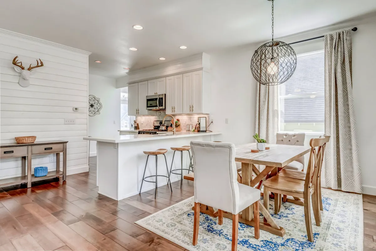 A wide interior shot capturing the open-plan dining and kitchen area. The image features a large wooden dining table set for a meal, a stylish kitchen island with seating, decorative bohemian light fixtures, and an accent wall that enhances the modern coastal aesthetic. Soft natural light streams in, creating a welcoming atmosphere ideal for social gatherings and family meals.