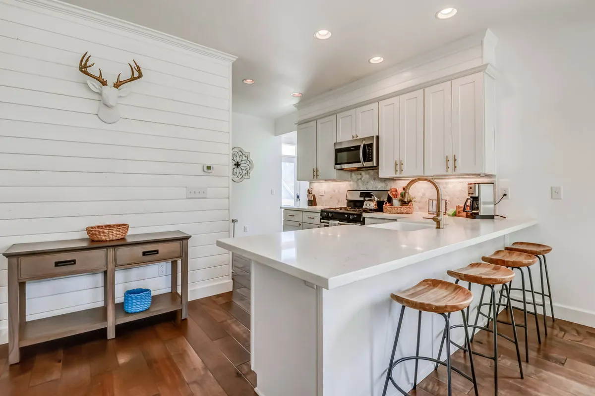 A stylish modern kitchen with a spacious island, sleek bar stools, white cabinetry, and eye-catching wooden accents. Unique wall decor, including a deer head, adds personality and a photo-ready backdrop. The space is both functional and visually appealing, perfect for entertaining or capturing content.