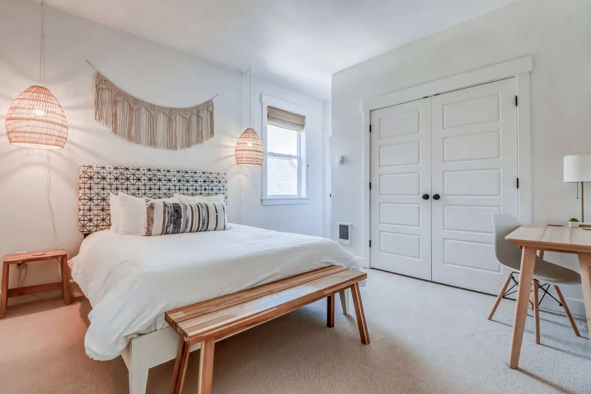 A peaceful bedroom featuring a bed with a patterned headboard, woven wall hanging, stylish pendant lights, a wooden bench at the foot of the bed, and a dedicated workspace with a desk and chair by the window. Soft natural light bathes the room, accentuating the cozy ambiance and designer décor.