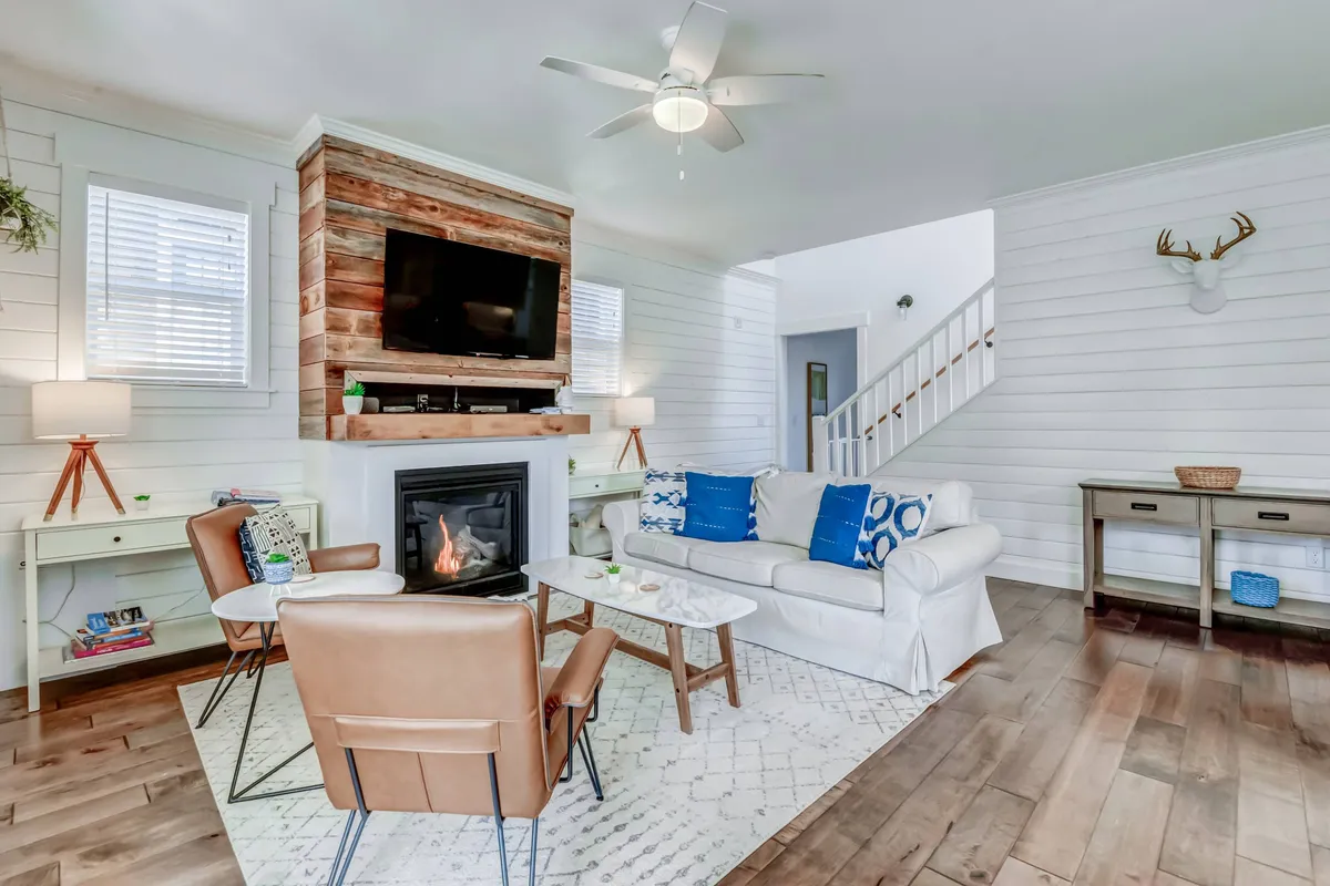 A cozy living room with a plush sofa, a prominent stone fireplace, a rustic wooden accent wall, and decorative antlers above the mantel. The room is bathed in natural light, creating an inviting setting for relaxing, entertaining, or unwinding after a day at the beach.